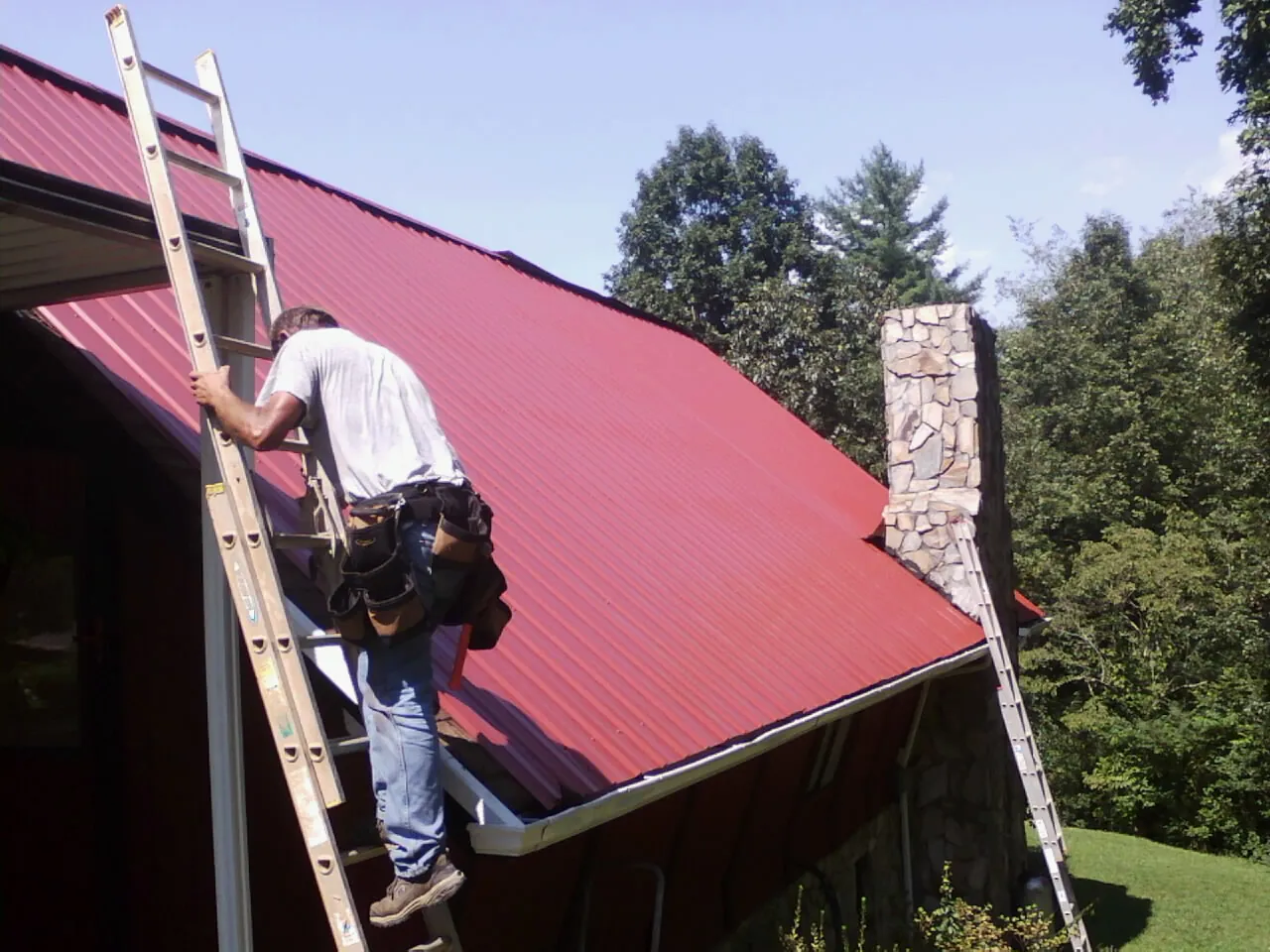 Roofer on a ladder beside a red metal roof and stone chimney — Soffit & Fascia Repair by Roofing Brothers in Cedar Lane